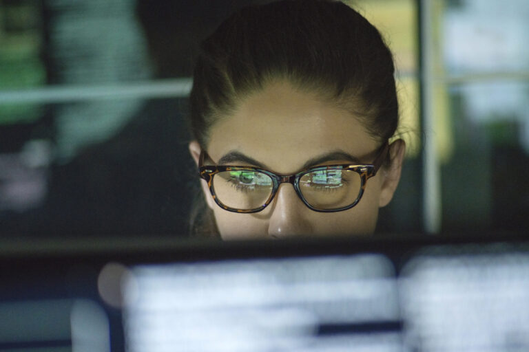 Close up of a young woman surrounded by monitors & their reflections displaying scrolling text and data.